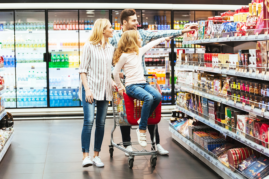 Cheery little girl sitting on a shopping cart and choosing candy with her parents at the supermarket